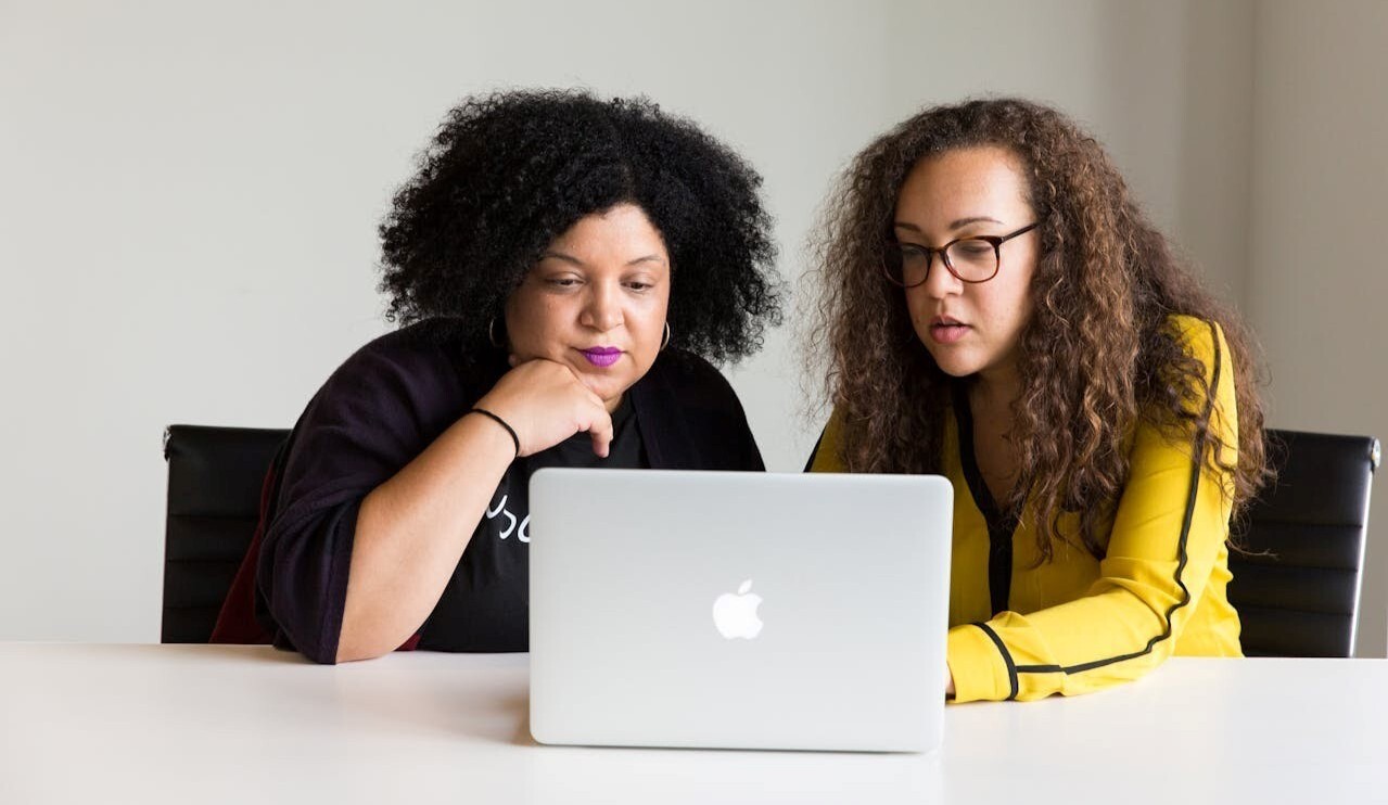 Twee vrouwen met krullend haar zitten achter een laptop die op tafel staat.