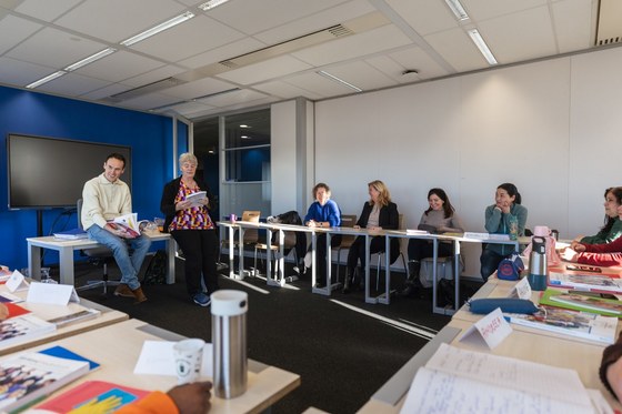 Er zitten mensen in een klaslokaal achter aan elkaar geschoven bureaus. Wethouder Paul Smeulders zit op de tafel en luistert naar de mensen in het lokaal.