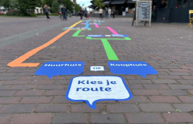 Gekleurde lijnen en tekstballonnen geplakt op het looppad naast Musis. Op de voorgrond staat 'Kies je route', gevolgd door 'Huurhuis' of 'Koophuis' als keuze.