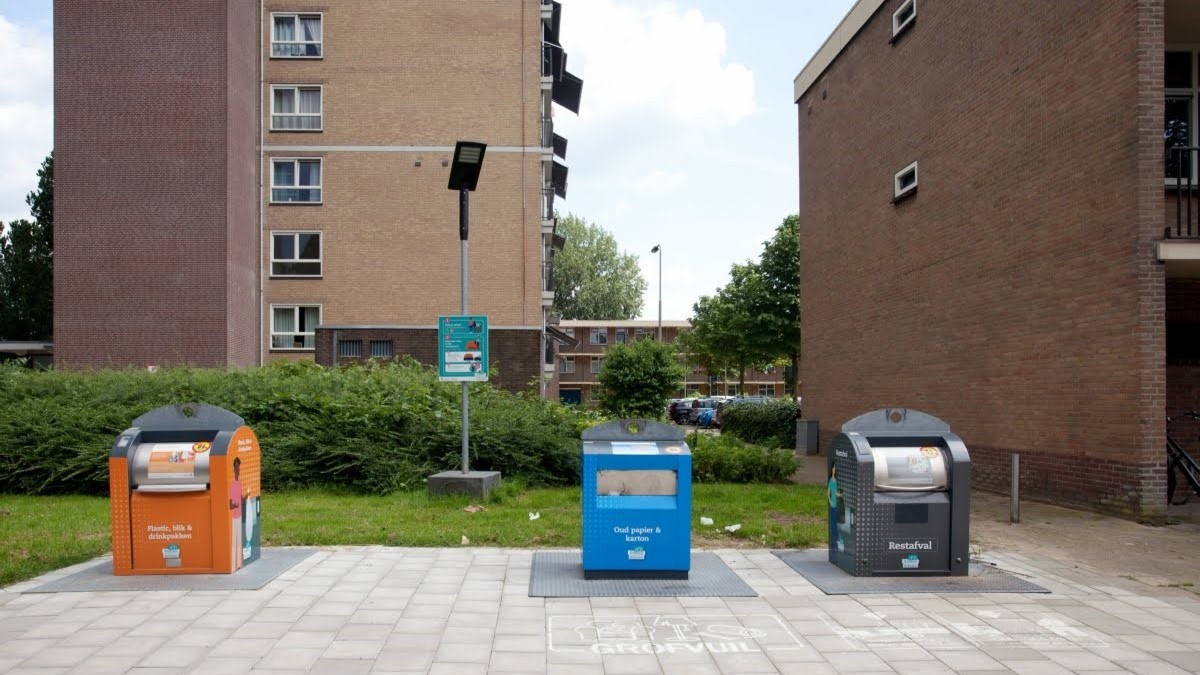 Een appartementencomplex waar drie ondergrondse containers voor staan: een oranje, een blauwe en een grijze.